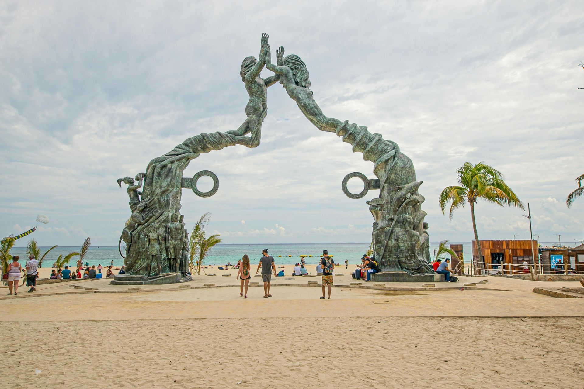 Arco escultórico en una playa de arena; figuras que se extienden hacia arriba, con el océano y la gente en el fondo.