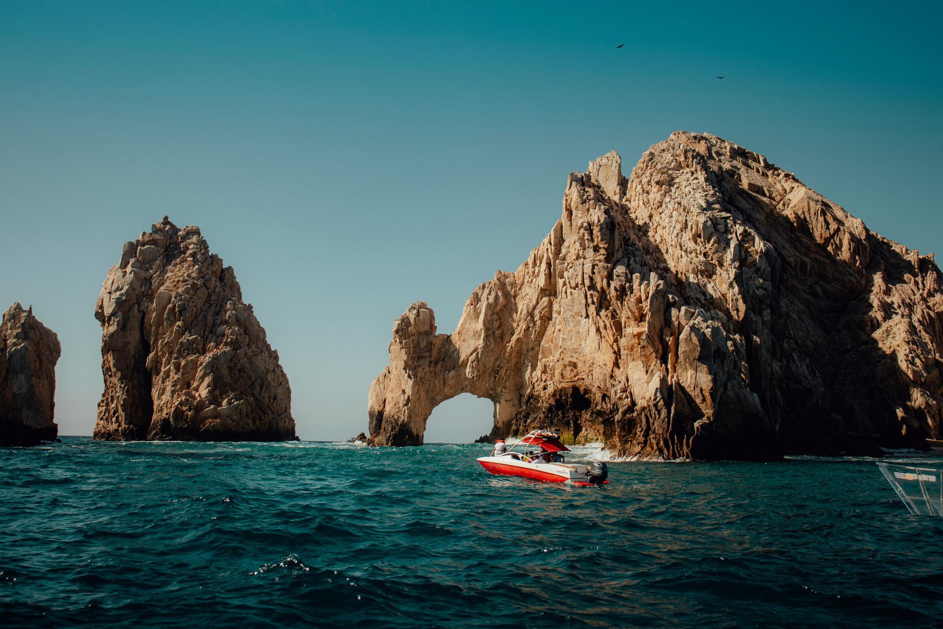 Acantilados rocosos se elevan desde el océano, con un barco rojo y blanco cerca del arco y un cielo azul claro.