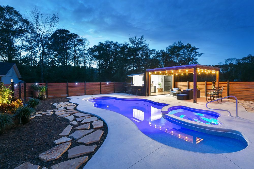 Nighttime backyard with a freeform pool, hot tub, and cabana with string lights; a wooden fence.