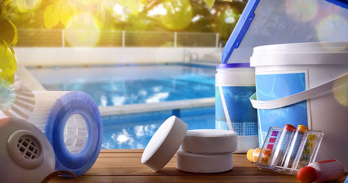 Pool maintenance supplies on a wooden surface with a pool in the background: filters, tablets, chemicals.