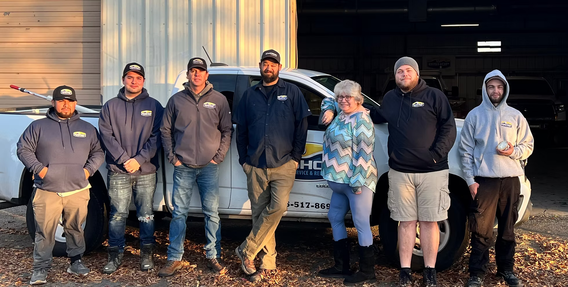 Group of people posing by a white truck with logo, outdoors. Several wear hoodies. Sunny day, dry leaves on the ground.