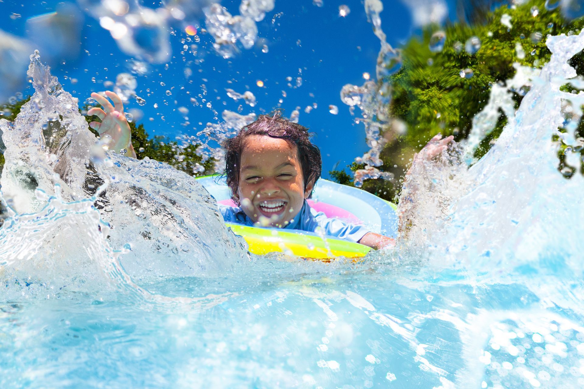 Boy in a pool with a flotation device, splashing water and smiling under a blue sky.