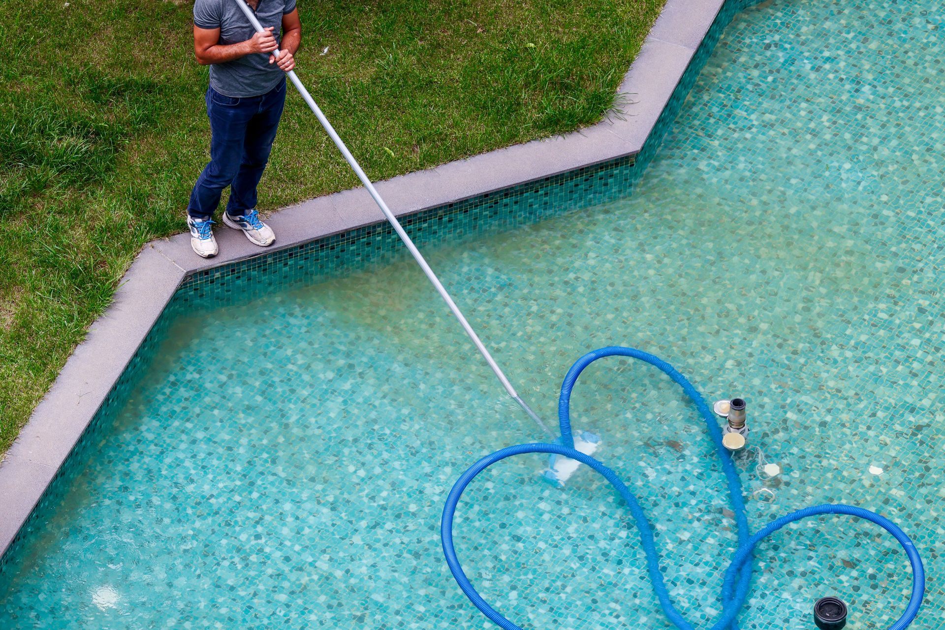 Person using a pool skimmer to clean a turquoise swimming pool with a blue hose