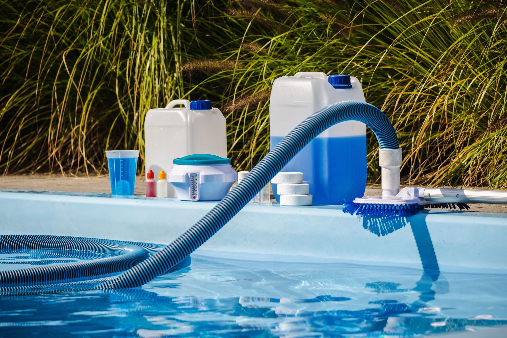 Pool cleaning equipment beside a blue swimming pool, with hoses, bottles, and containers on the deck.