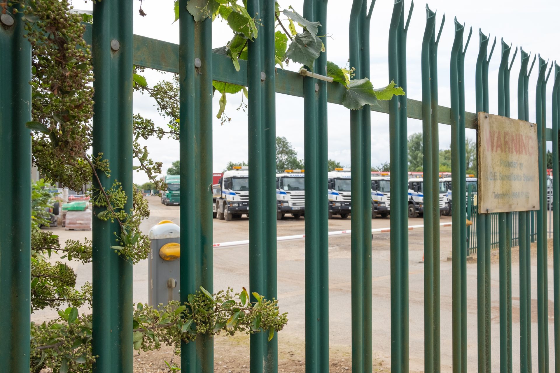 A green palisade fence with a warning sign guards a yard of fleet trucks.