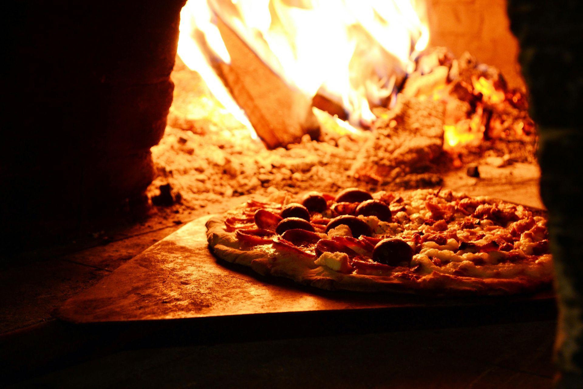 Una pizza condita con olive e peperoni rossi, cotta su una pala di legno all'interno di un forno a legna in mattoni.