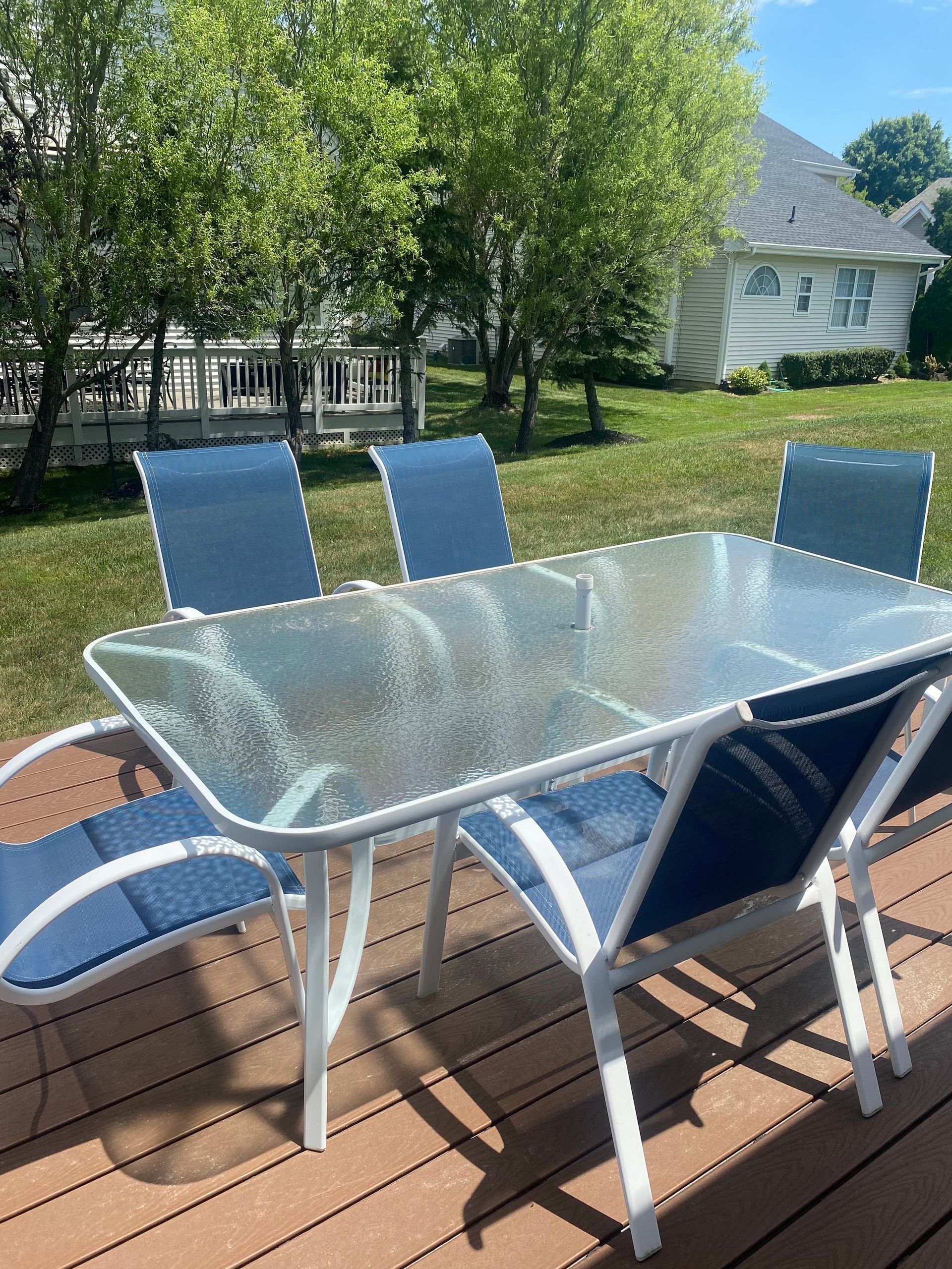 Outdoor glass-topped table and six blue chairs on a wooden deck in front of a green lawn and trees.