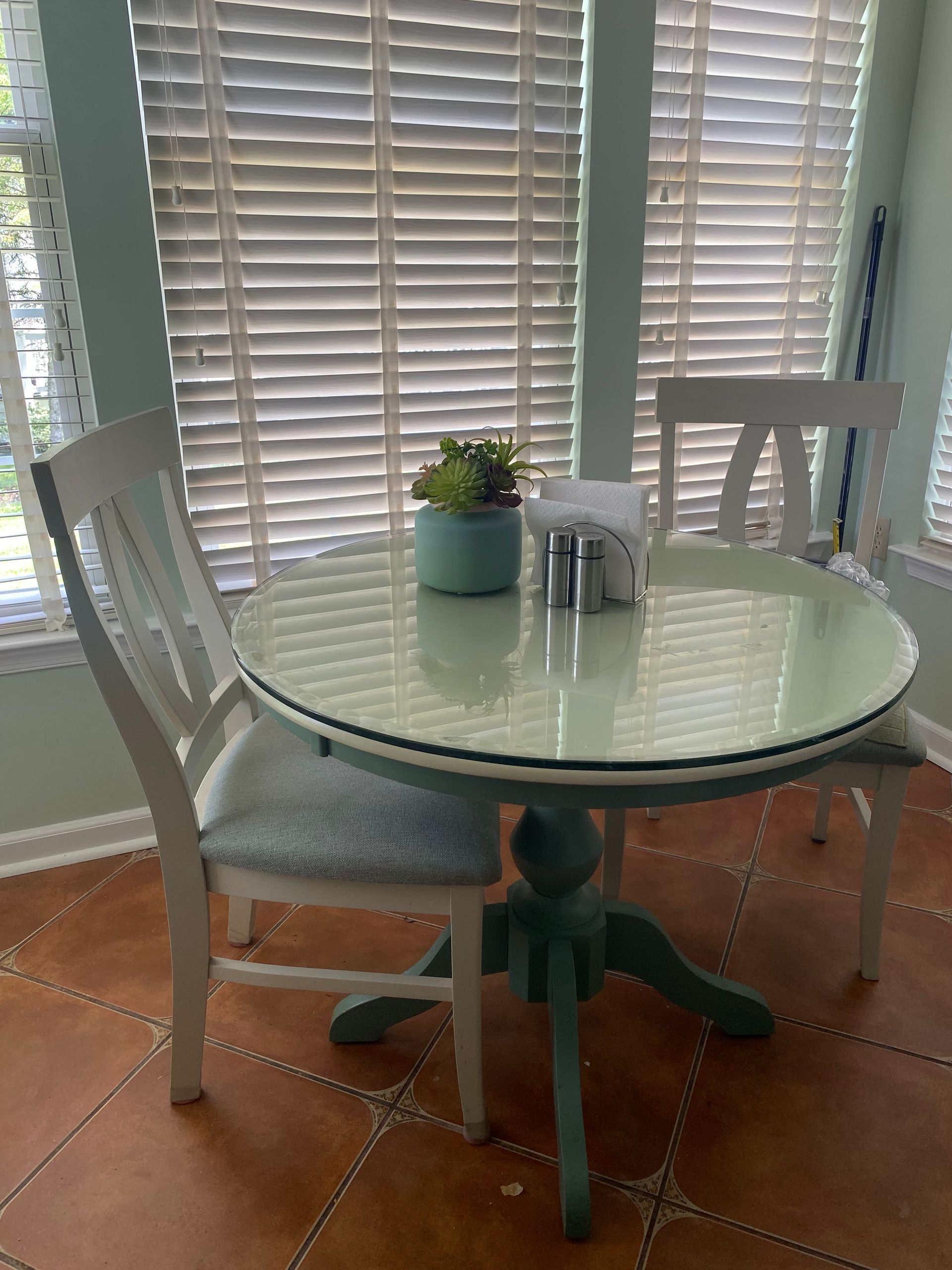 A small, round, mint-green table with two white chairs in front of a window with blinds.