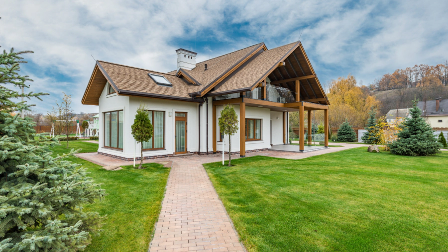 White house with a brown roof and a porch, set on a green lawn with trees and a paved walkway.