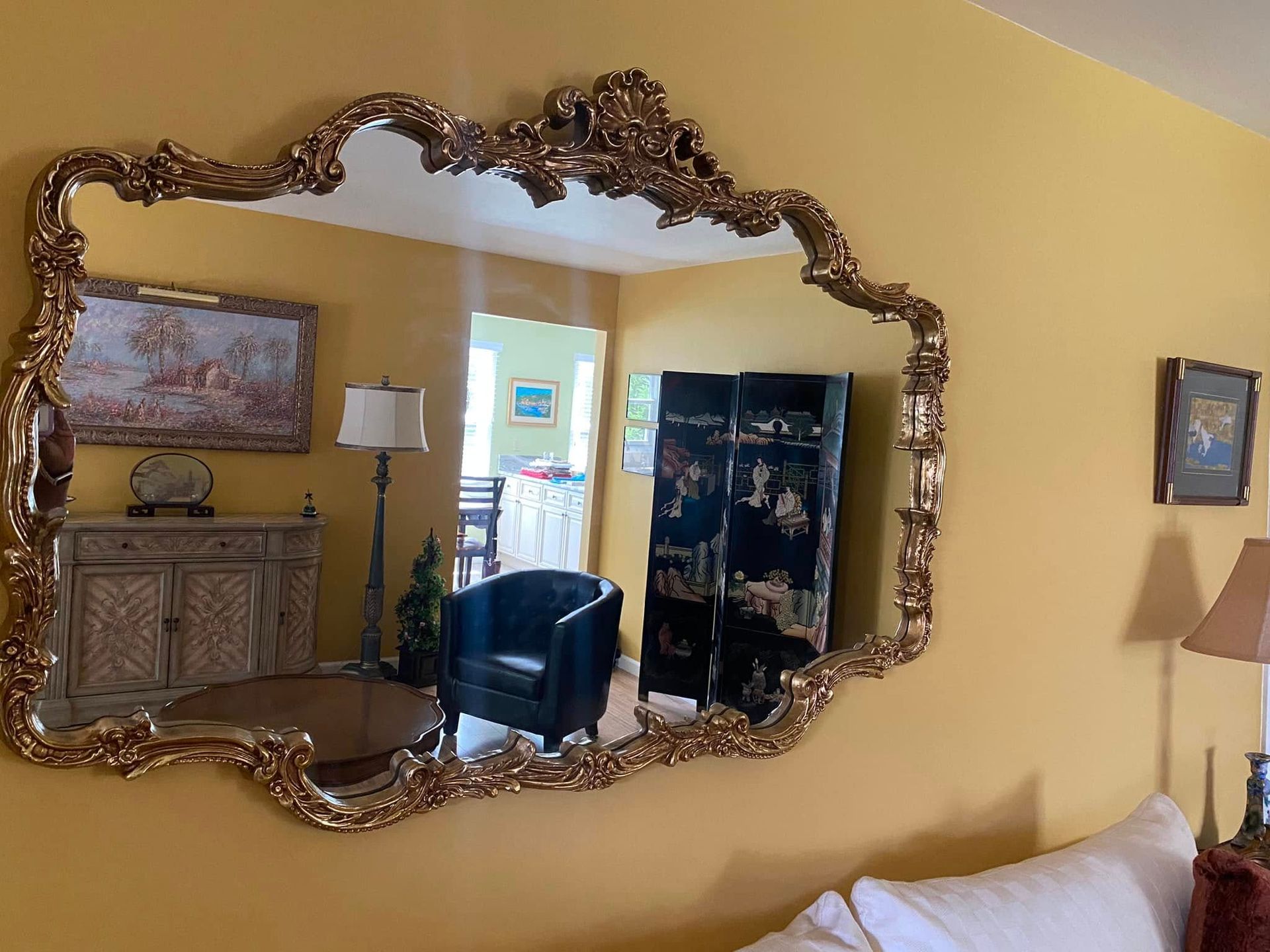 Large ornate gold-framed mirror on a yellow wall, reflecting a room with furniture and a folding screen.