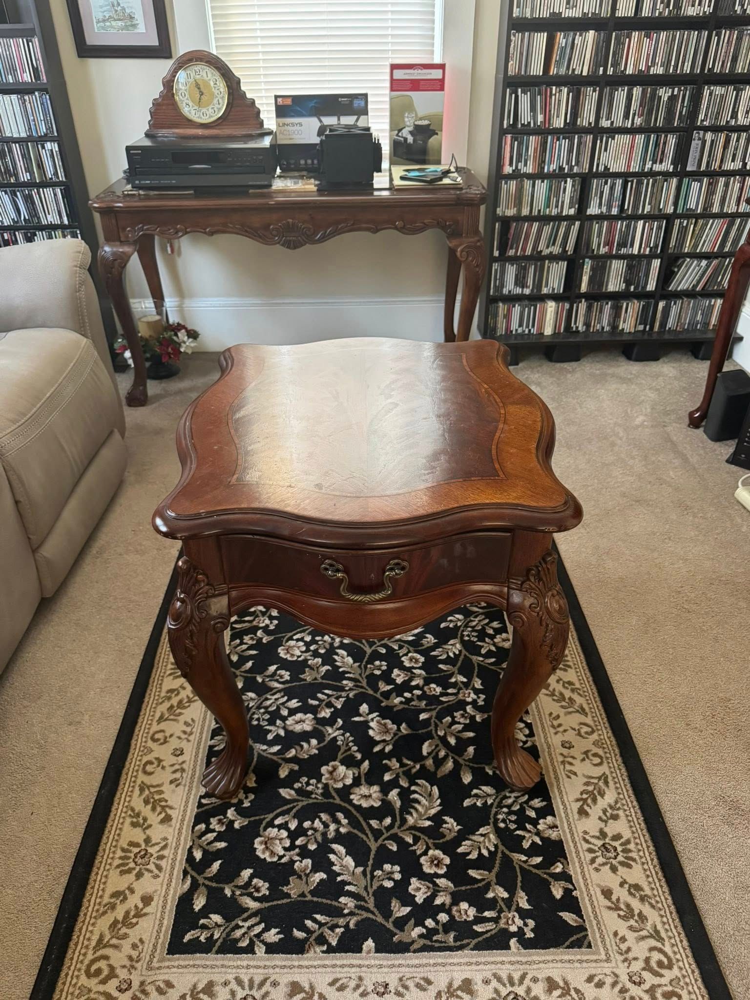 Wooden coffee table on floral rug, with an antique table and wall shelves in background.