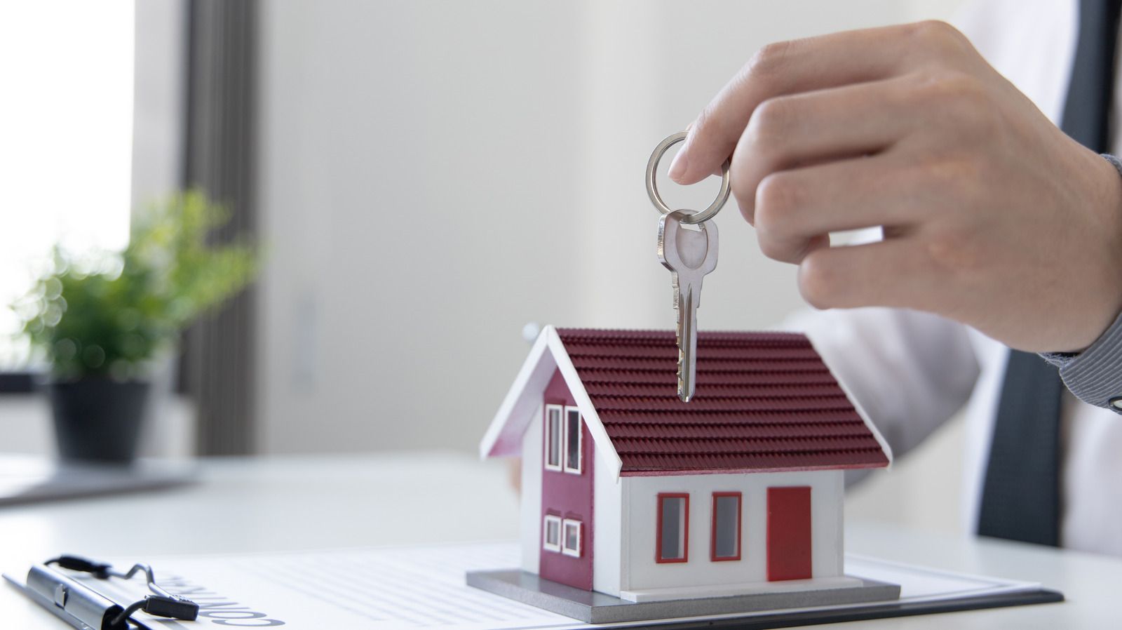 Hand holding house key over a miniature house, possibly symbolizing a real estate transaction.