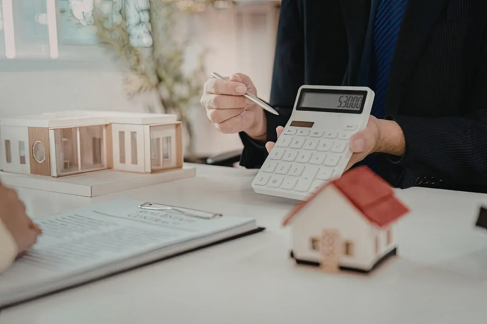 Person calculating with calculator, pointing at paperwork, model houses and red-roofed house on a desk.