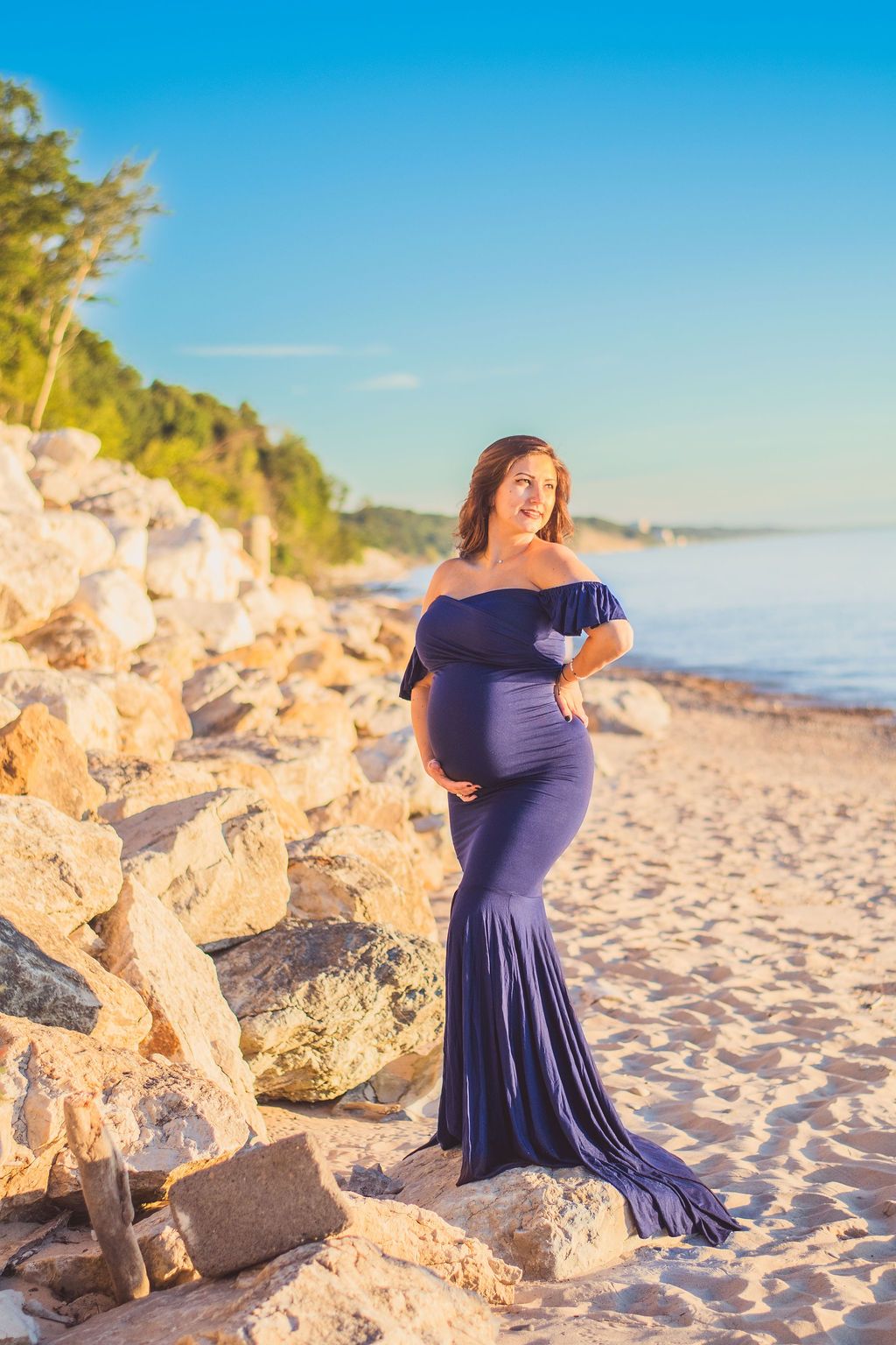 Pregnant woman in a navy gown standing on a sandy beach beside rocky cliffs at sunset