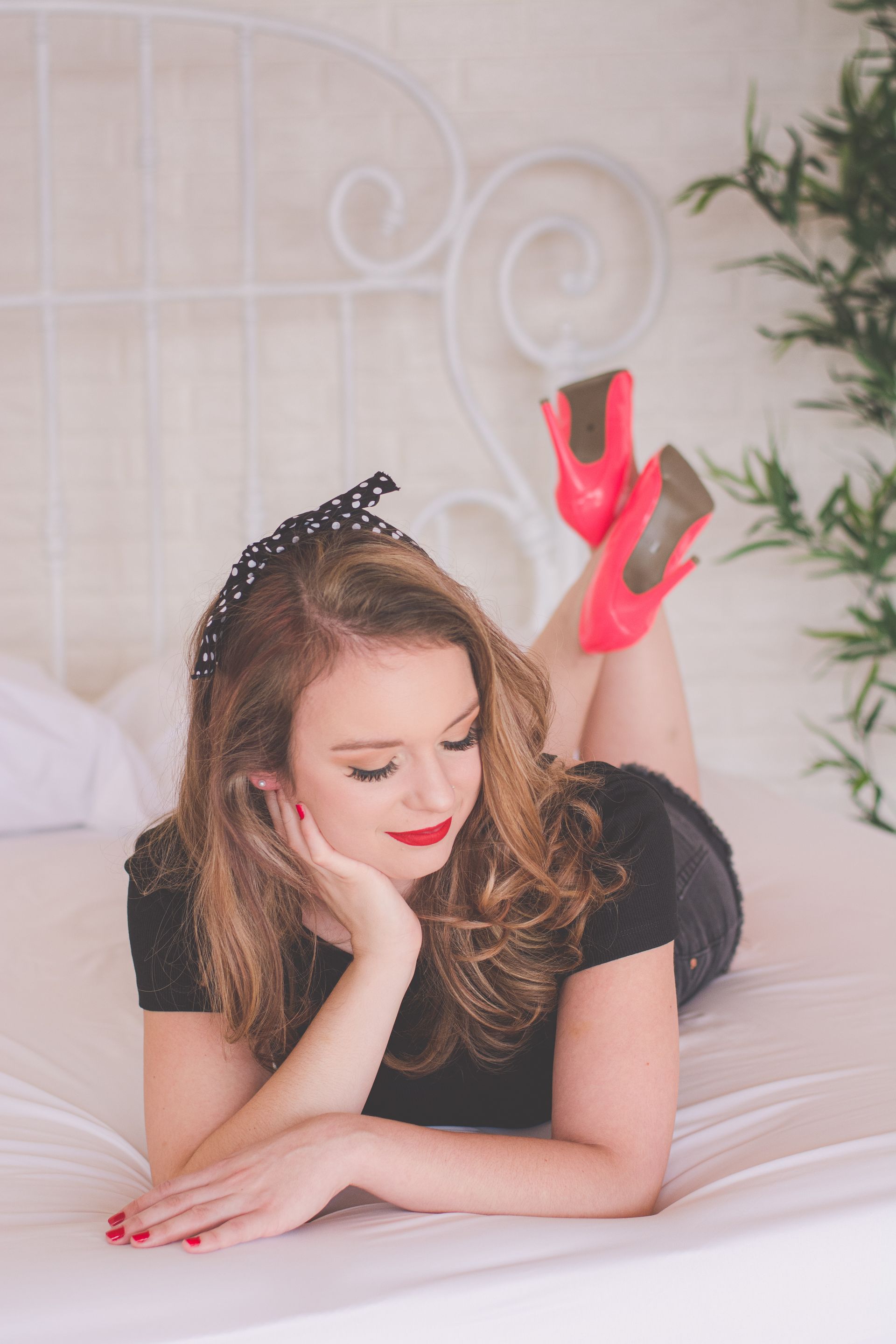 Woman in black dress lying on a bed, posing with bright pink heels and a black headband