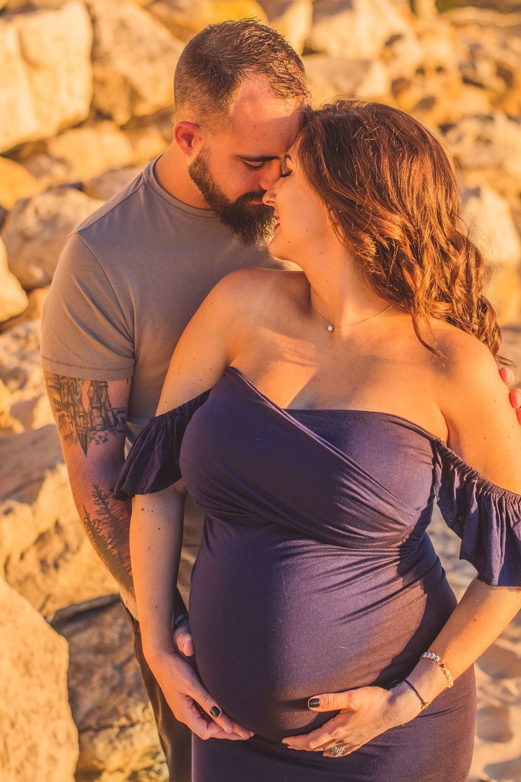 Couple embracing outdoors at sunset, pregnant woman in a blue dress holding her belly.