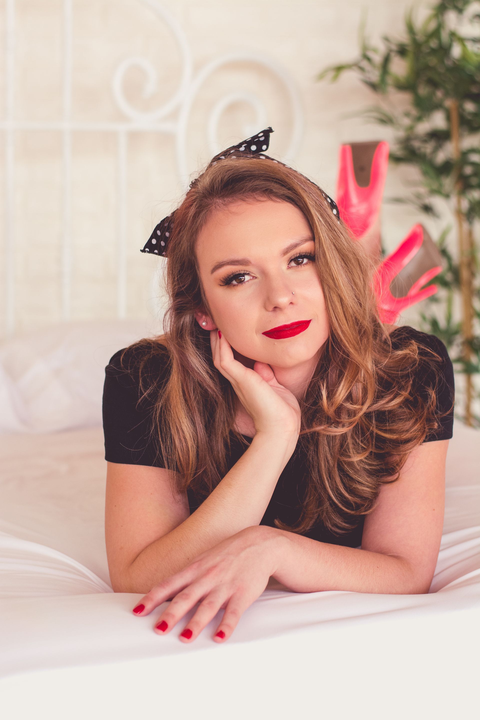 Woman lying on a bed with chin in hand, wearing a black top and red lipstick, in a soft pink bedroom.