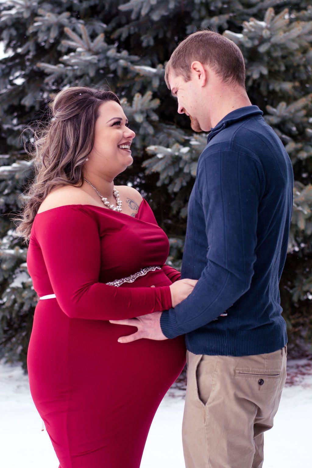 Couple standing in snow, smiling and holding hands, with evergreen trees in the background