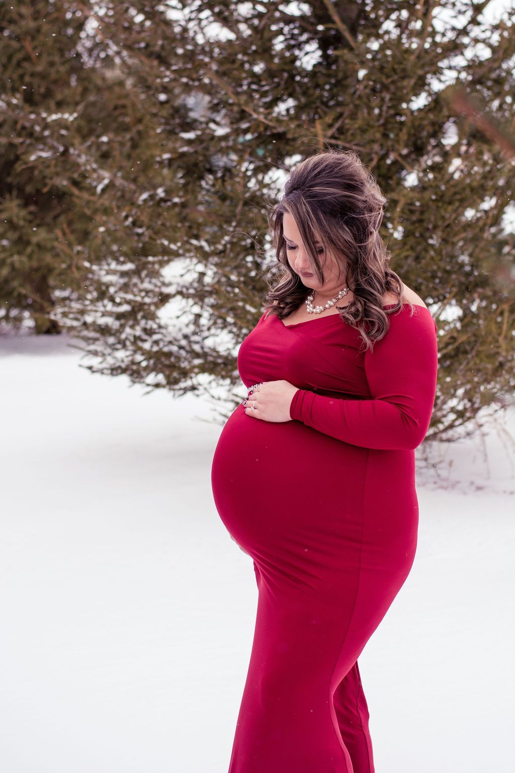 Pregnant person in a red dress standing in snow beside trees, looking down and holding their belly
