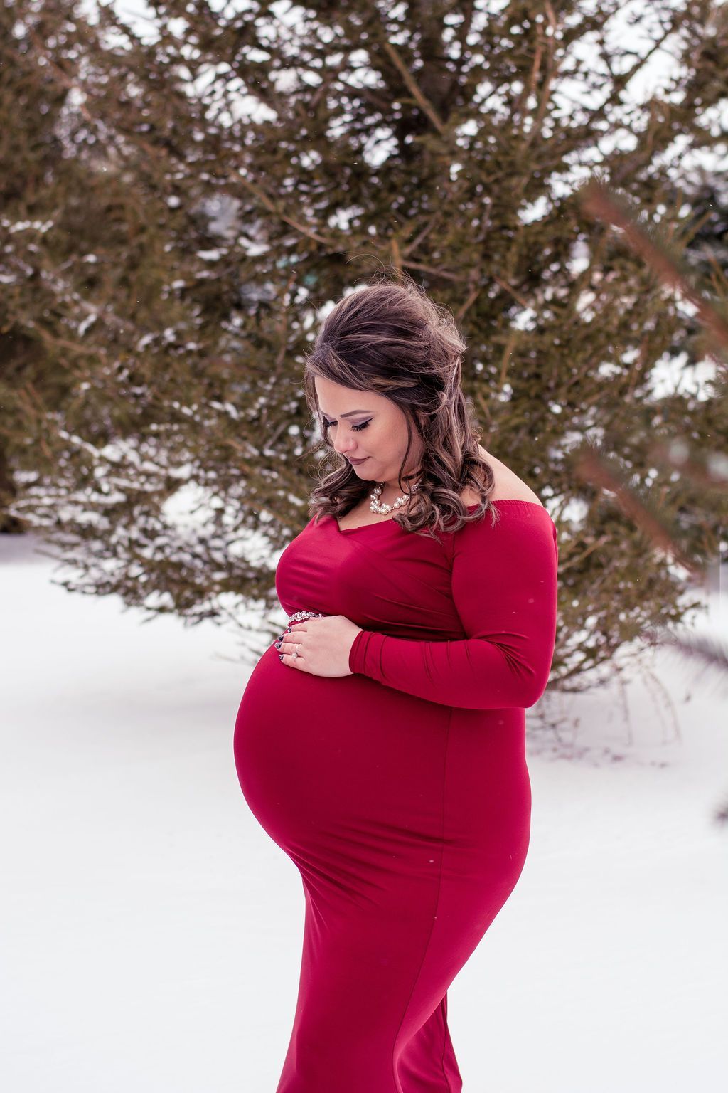 Pregnant person in a red dress standing in snow, looking down with hands on belly.