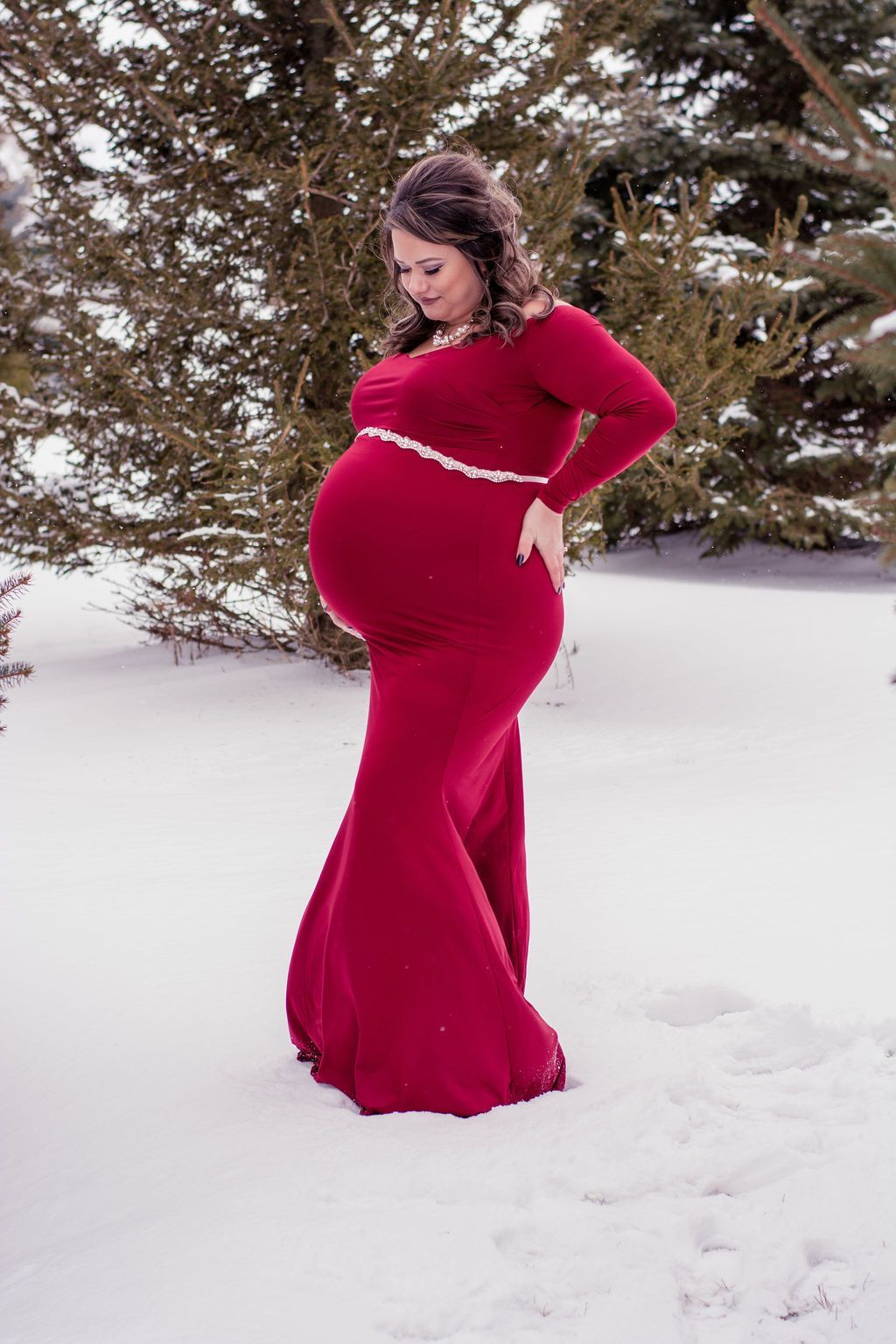 Pregnant woman in a red dress standing in snow by trees