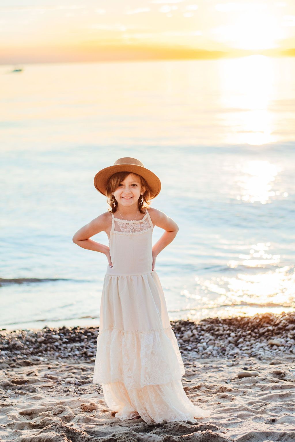 Child in a white dress and straw hat standing on a beach at sunset.