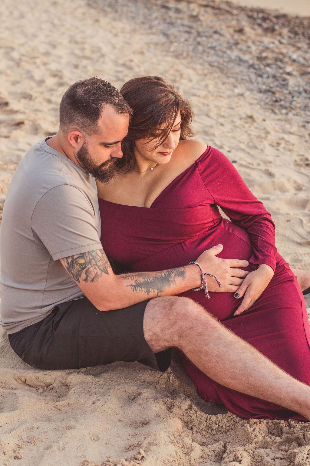 Couple sitting on sand, embracing as they look at a pregnant belly.