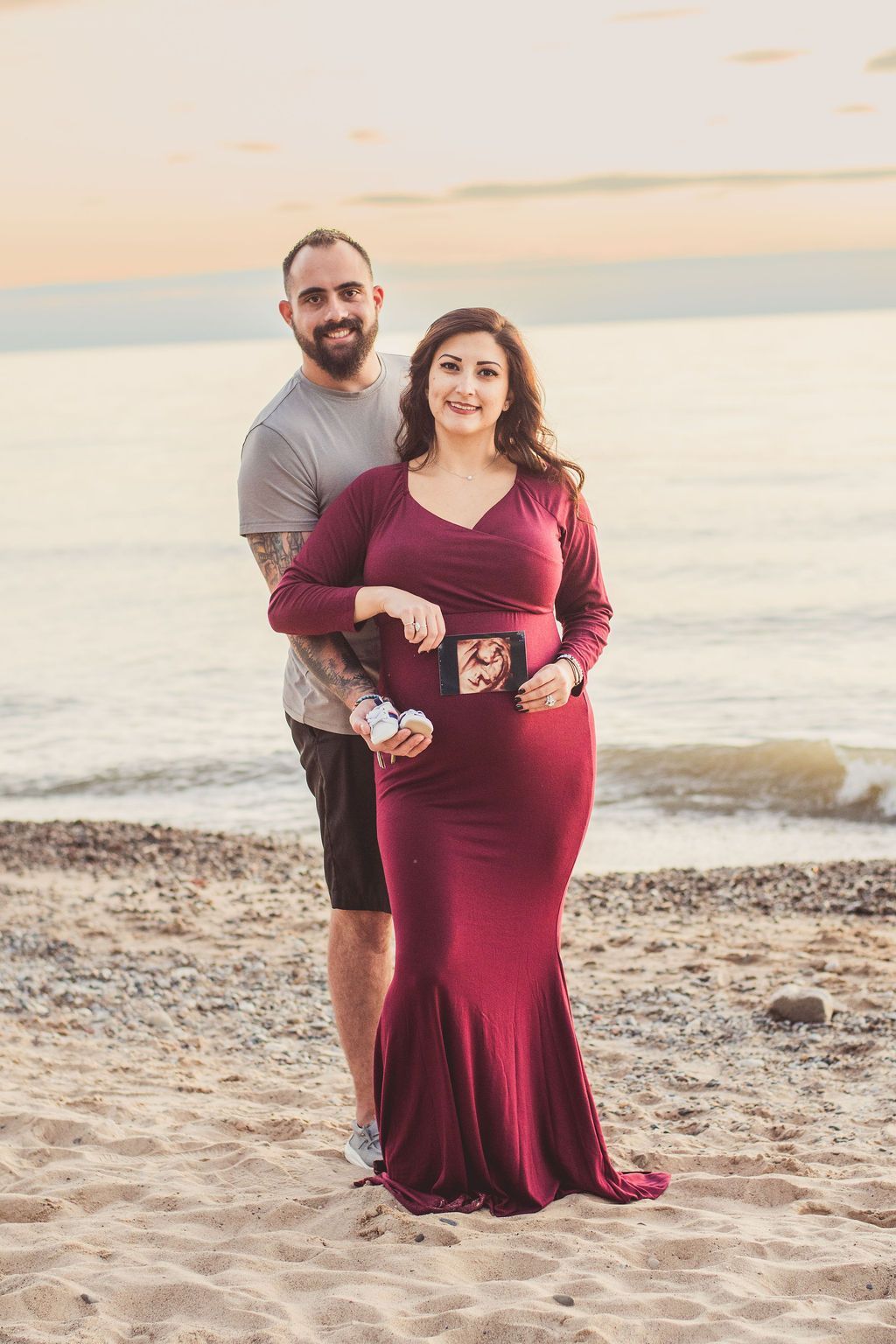 Couple standing on a beach at sunset, the woman in a burgundy dress holding a mug.