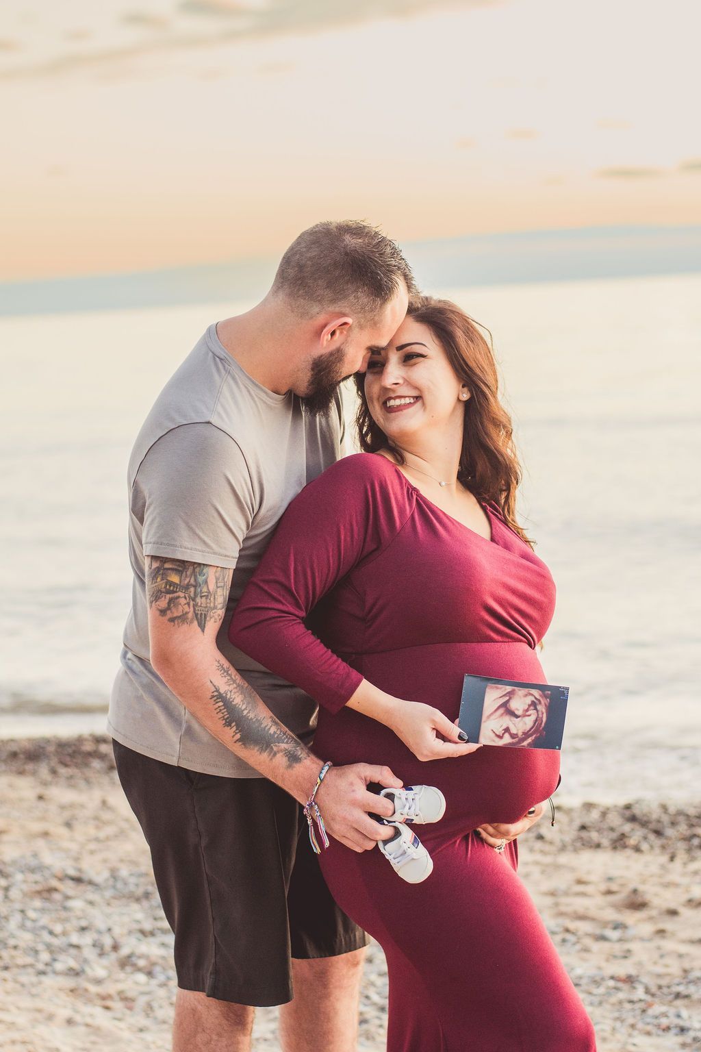 Pregnant woman in red dress on beach, smiling with partner at sunset, holding baby shoes and ultrasound photo