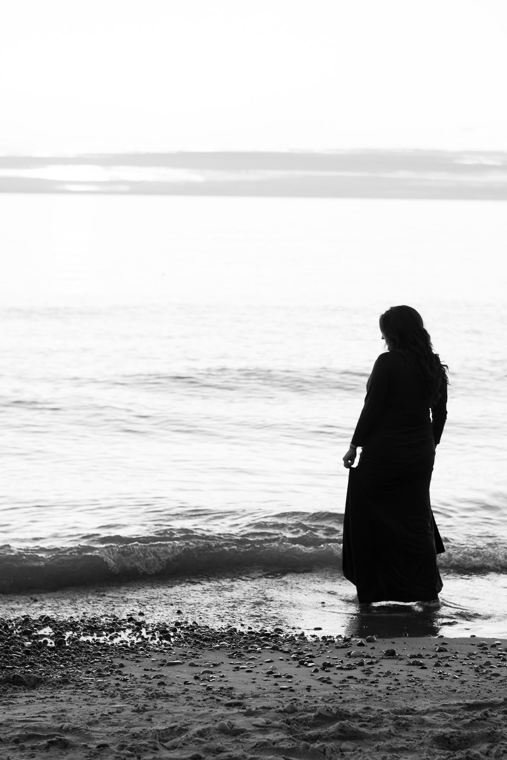 Silhouette of a person standing at the edge of a calm beach, facing the water at sunset.