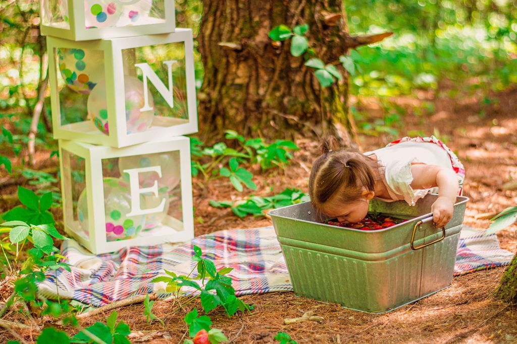 Child leaning into a green basket outdoors beside block letters spelling “ONE” under a tree
