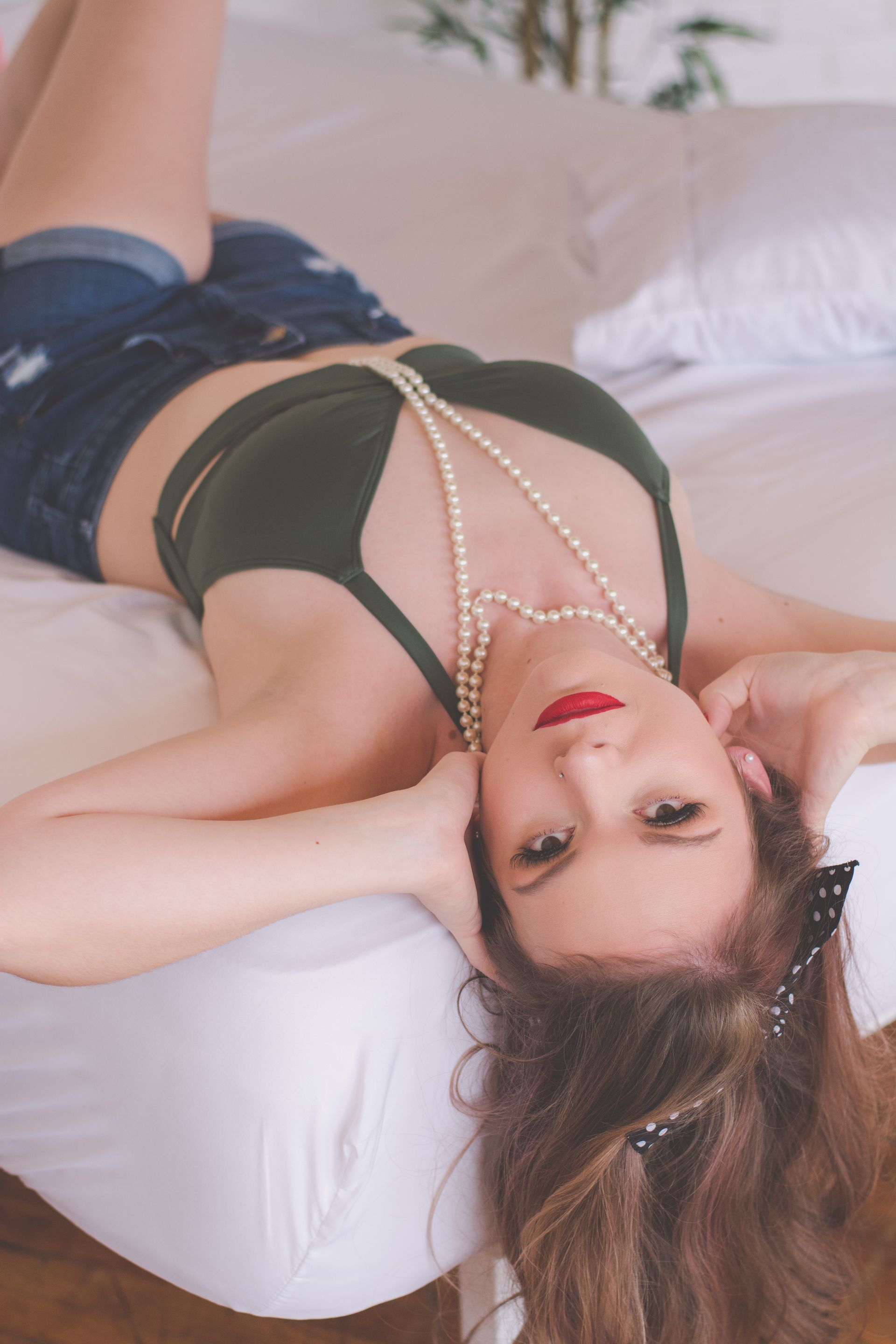 Woman reclining on a bed in a black top and pearl necklace, looking at the camera.