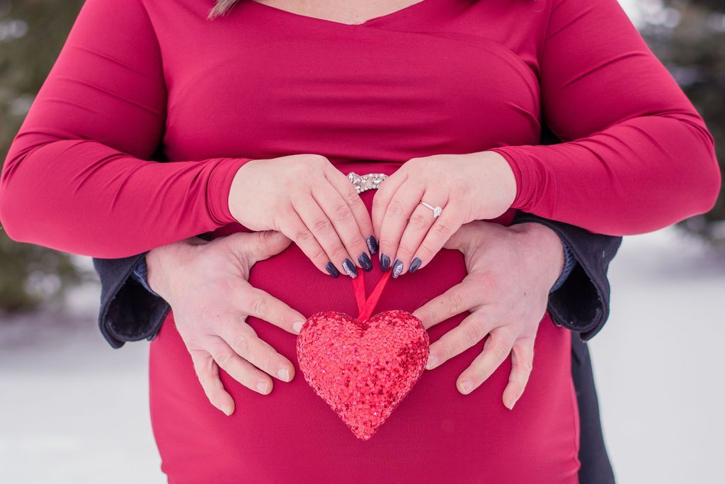 Hands forming a heart over a pink sweater, holding a small red heart pendant in the snow