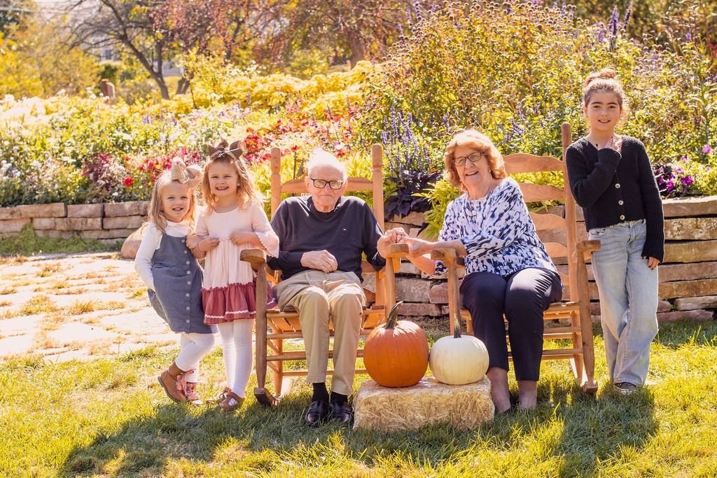 Family gathered outdoors in a sunny autumn garden, seated on chairs beside pumpkins and flowers.