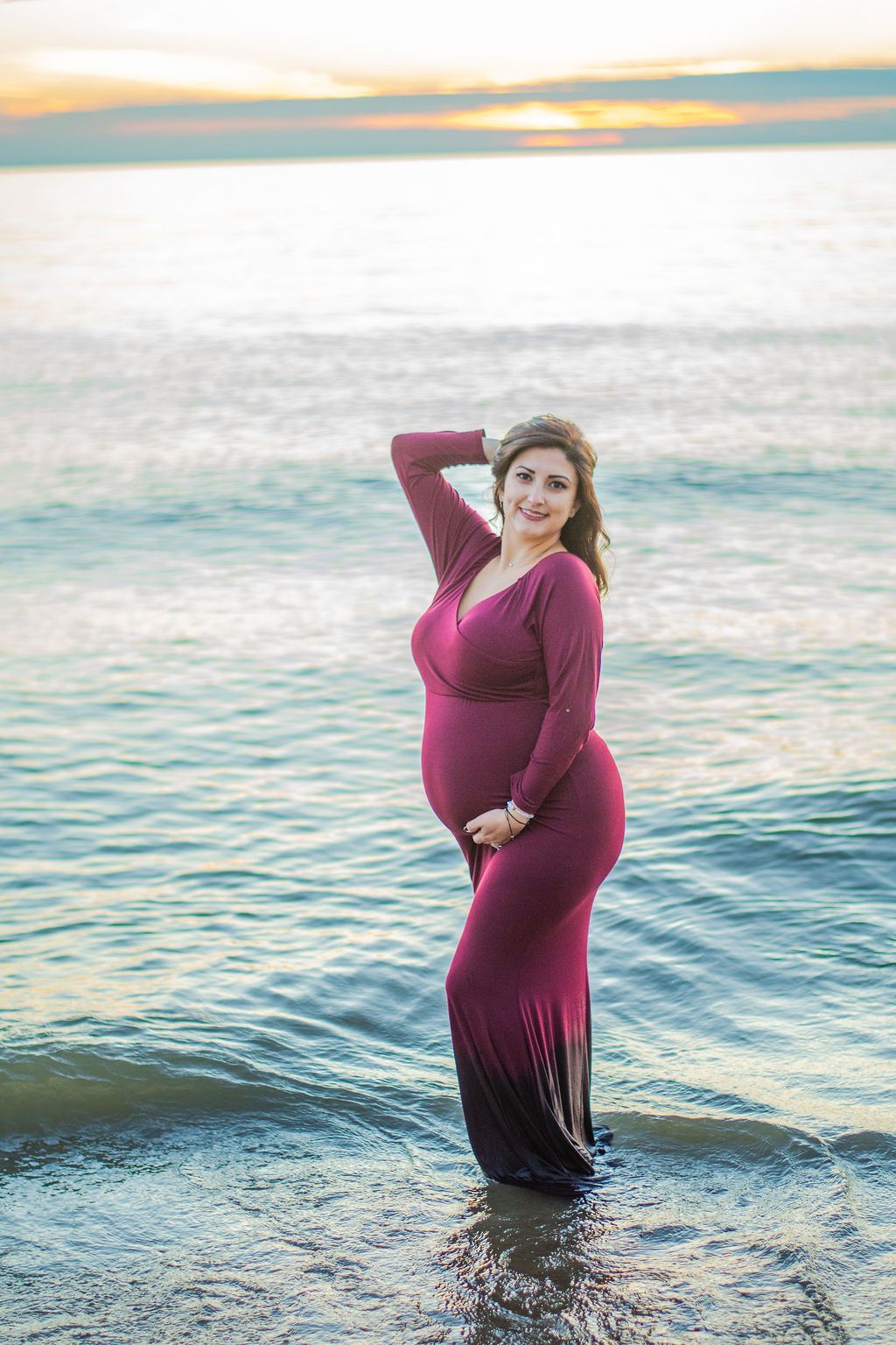 Pregnant woman in a magenta dress standing at the shoreline at sunset