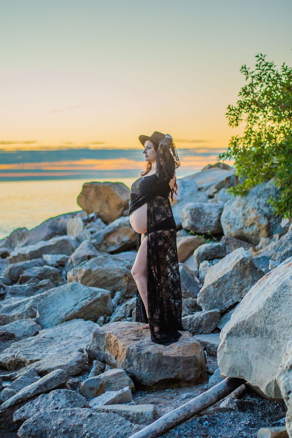 Person in a black dress standing on rocky shore at sunset, holding a bouquet by the water.