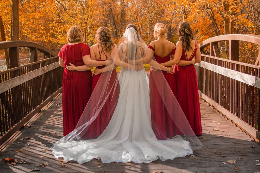 Bride and four bridesmaids on a wooden bridge in autumn, seen from behind, with flowing veils and red dresses