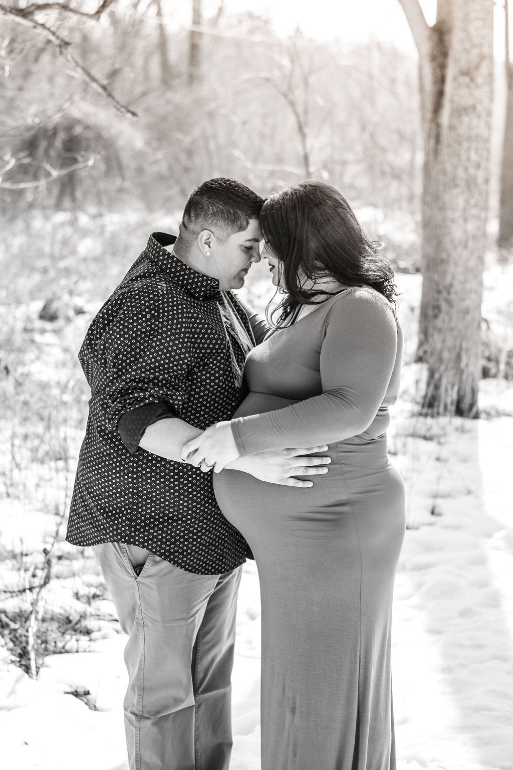 Couple embracing outdoors in snowy woods, touching foreheads with a large belly visible