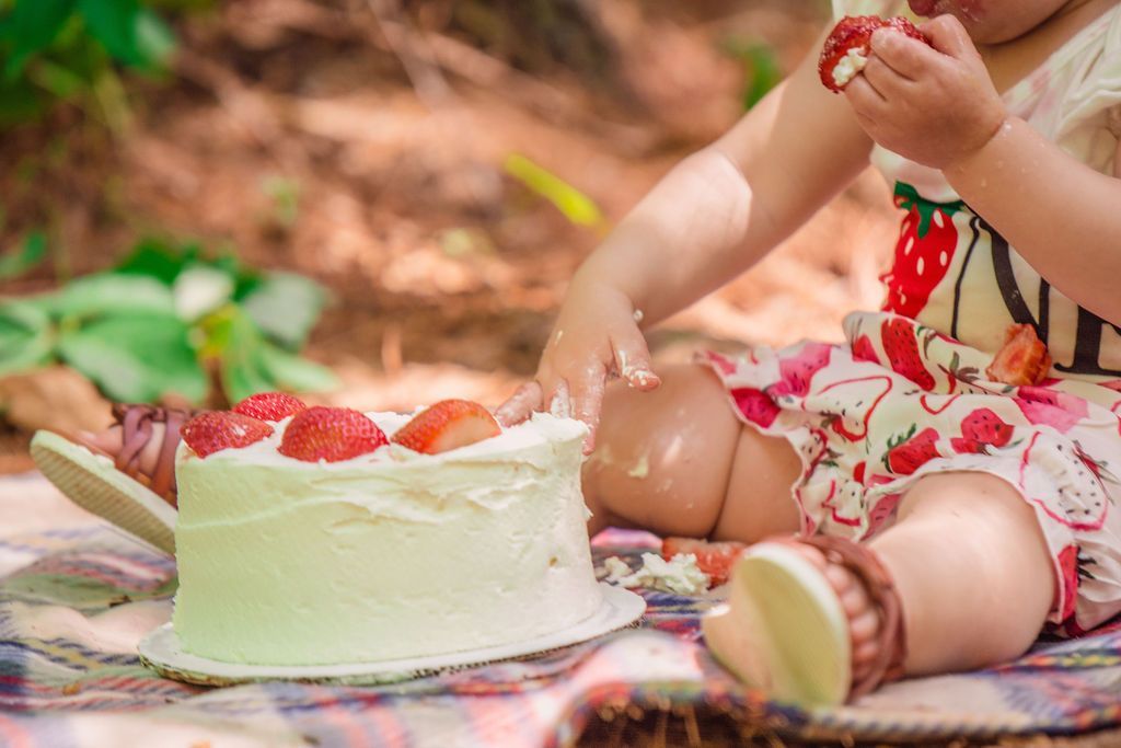 Child in a floral dress eating cake at a picnic blanket outdoors with strawberries.