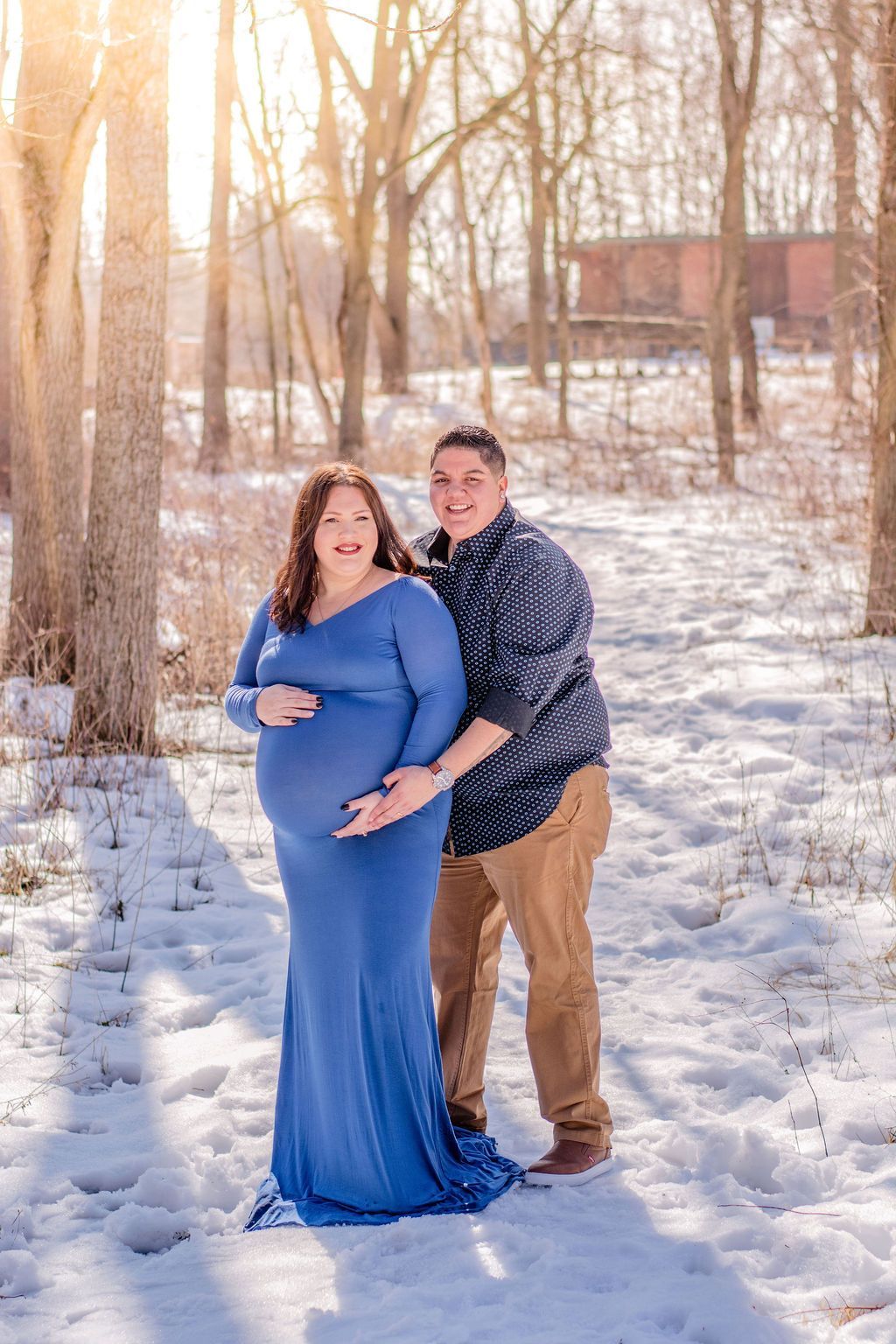 Couple standing in a snowy forest at sunset, with the woman in a blue dress and the man behind her.