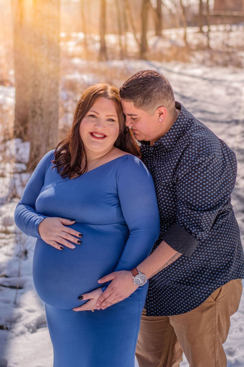 Couple embracing outdoors in winter, with the woman in a blue dress showing a pregnant belly.