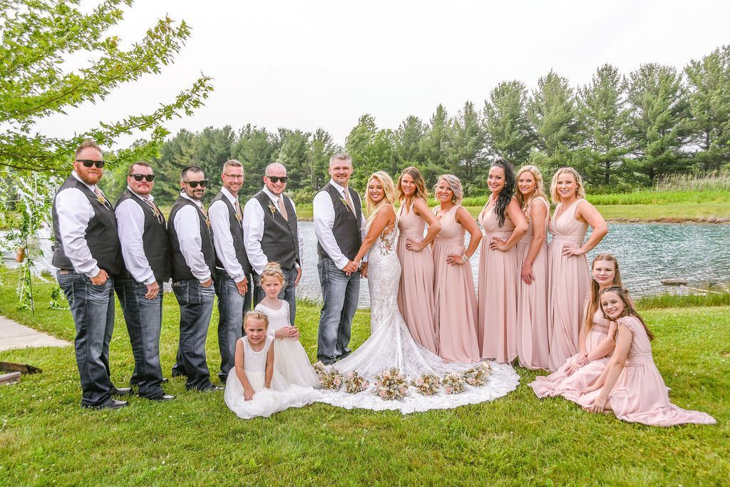 Wedding party posing outdoors beside a pond, with bride, groom, bridesmaids, groomsmen, and flower girls.