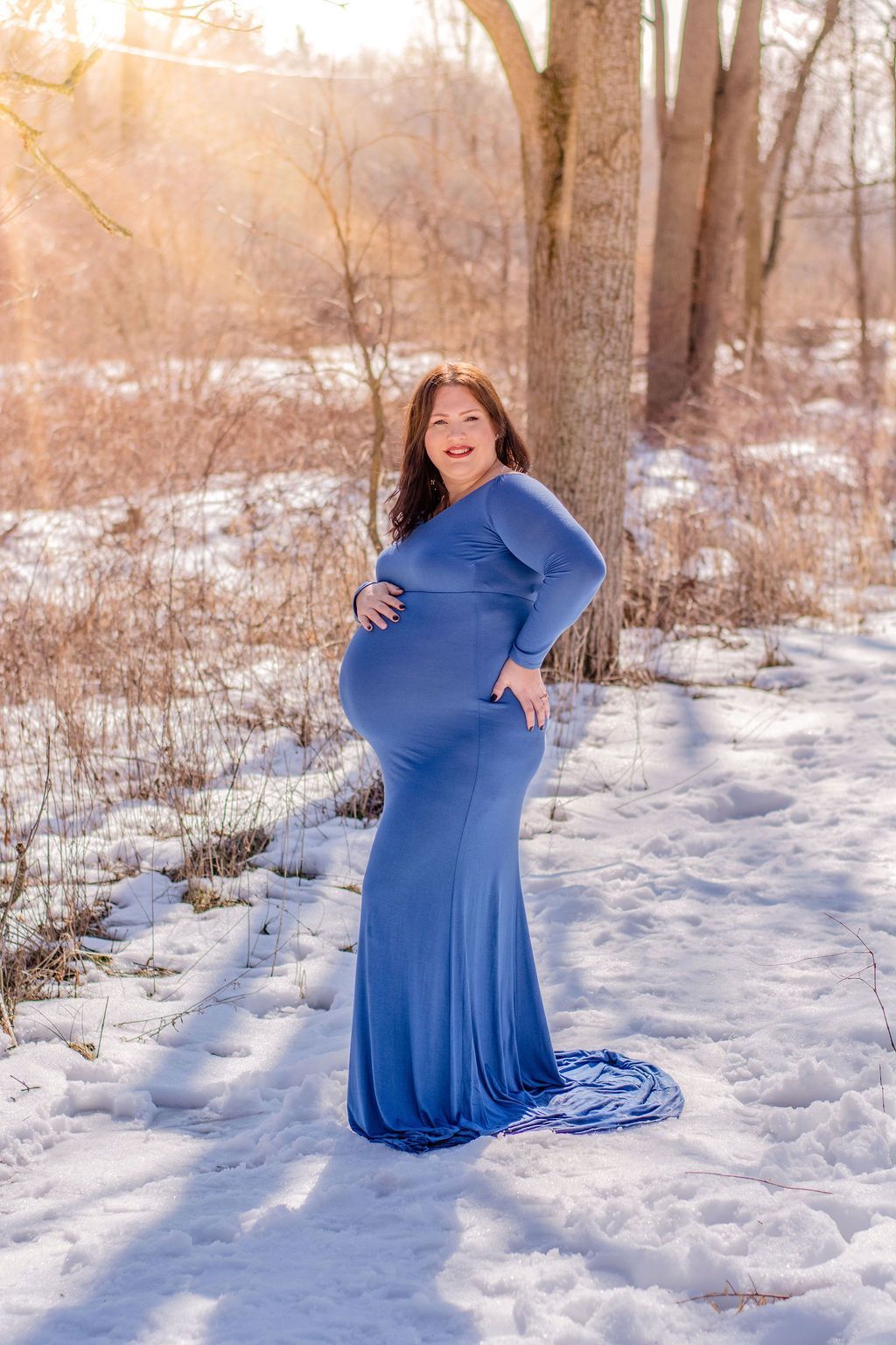Pregnant woman in a blue dress standing on a snowy path in a winter woodland