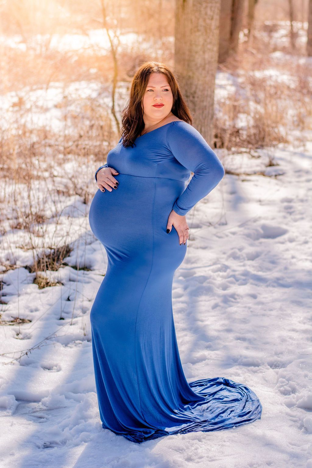 Pregnant person in a blue dress posing outdoors on a snowy path with trees in the background