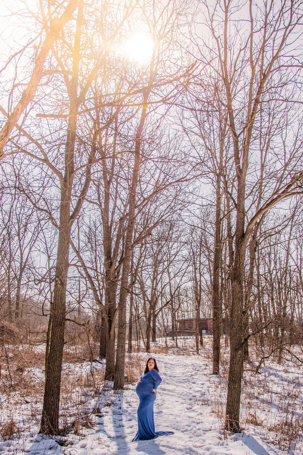 Person in a blue coat standing on a snowy path in a leafless forest under bright sun.
