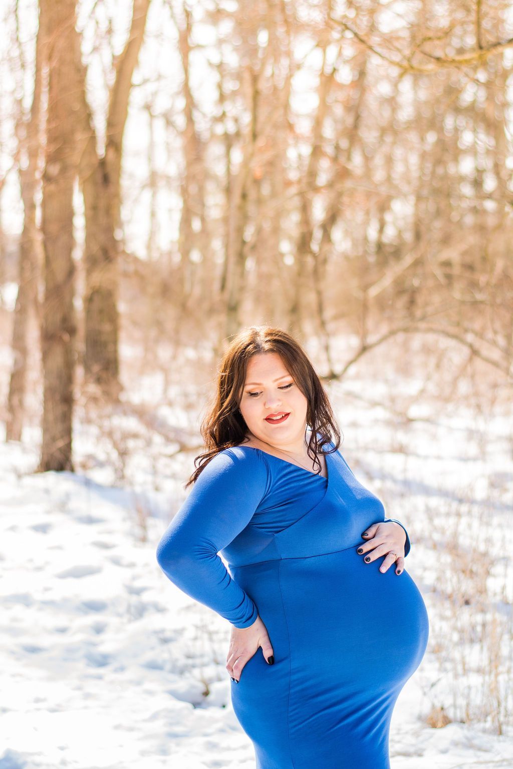 Pregnant woman in a blue dress standing in a snowy forest, smiling at the camera