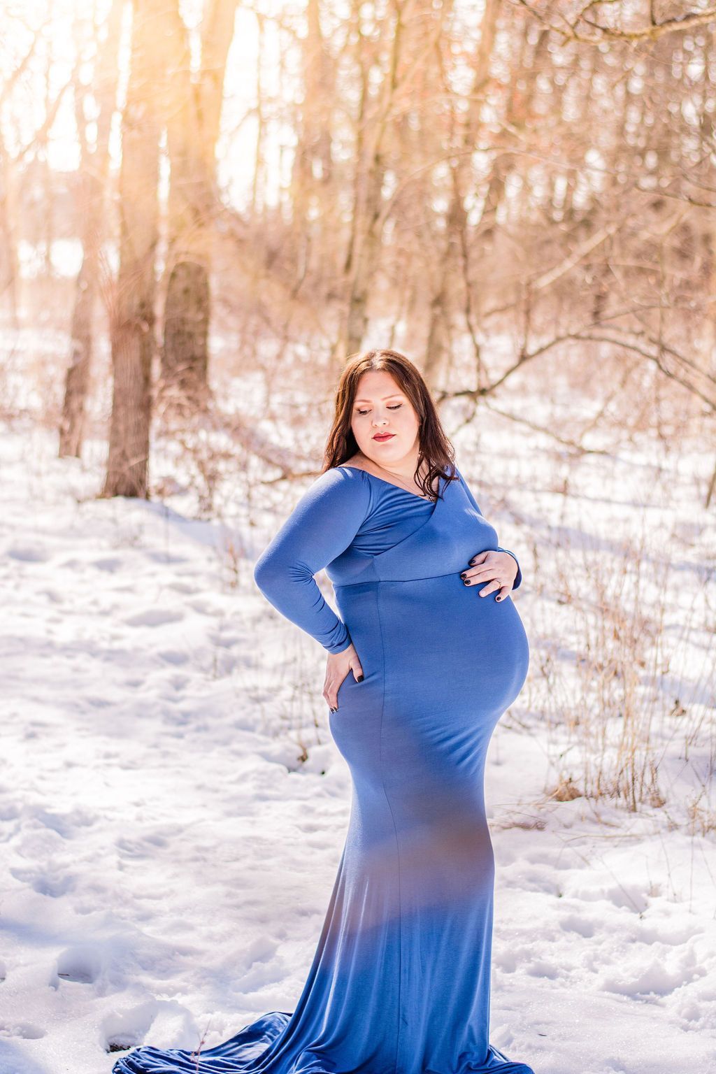 Pregnant person in a long blue dress standing in a snowy forest