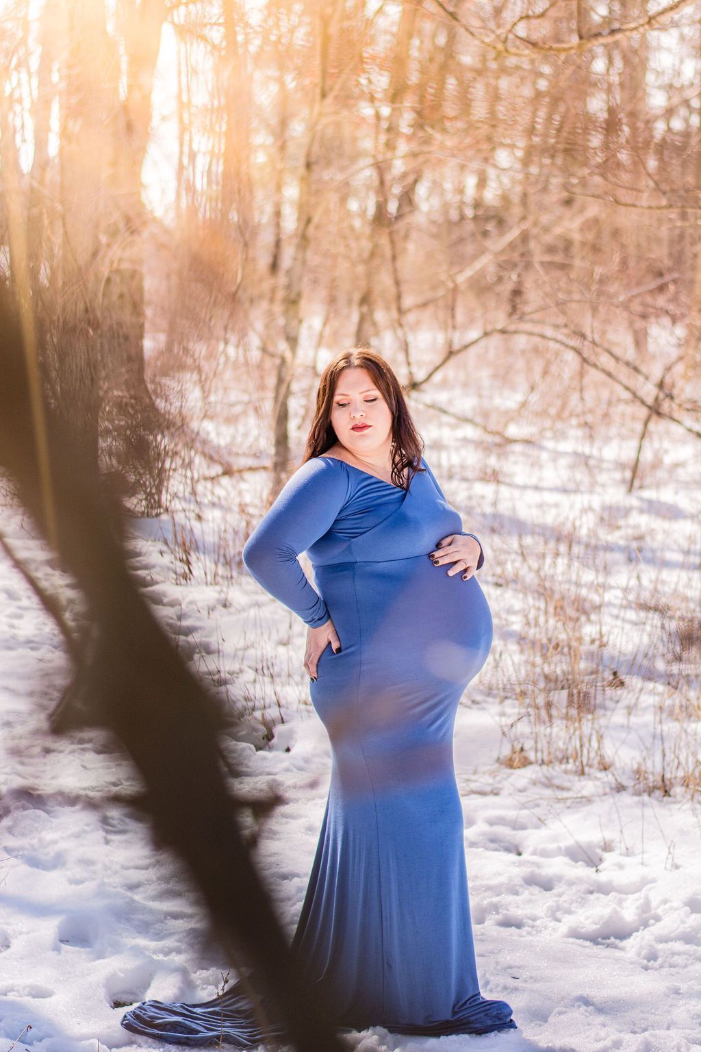 Pregnant woman in a blue dress standing in a snowy woodland, facing the camera