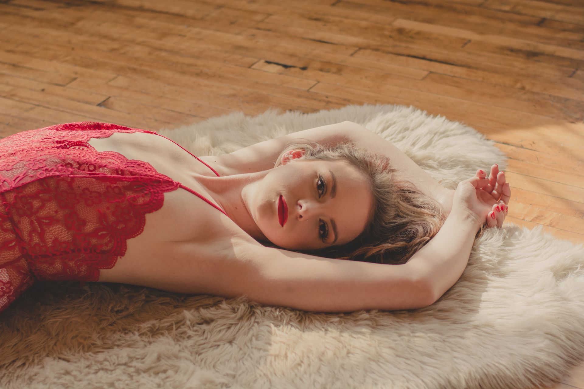 Person in a red lace dress lying on a white fur rug on a wooden floor, arms raised above head.