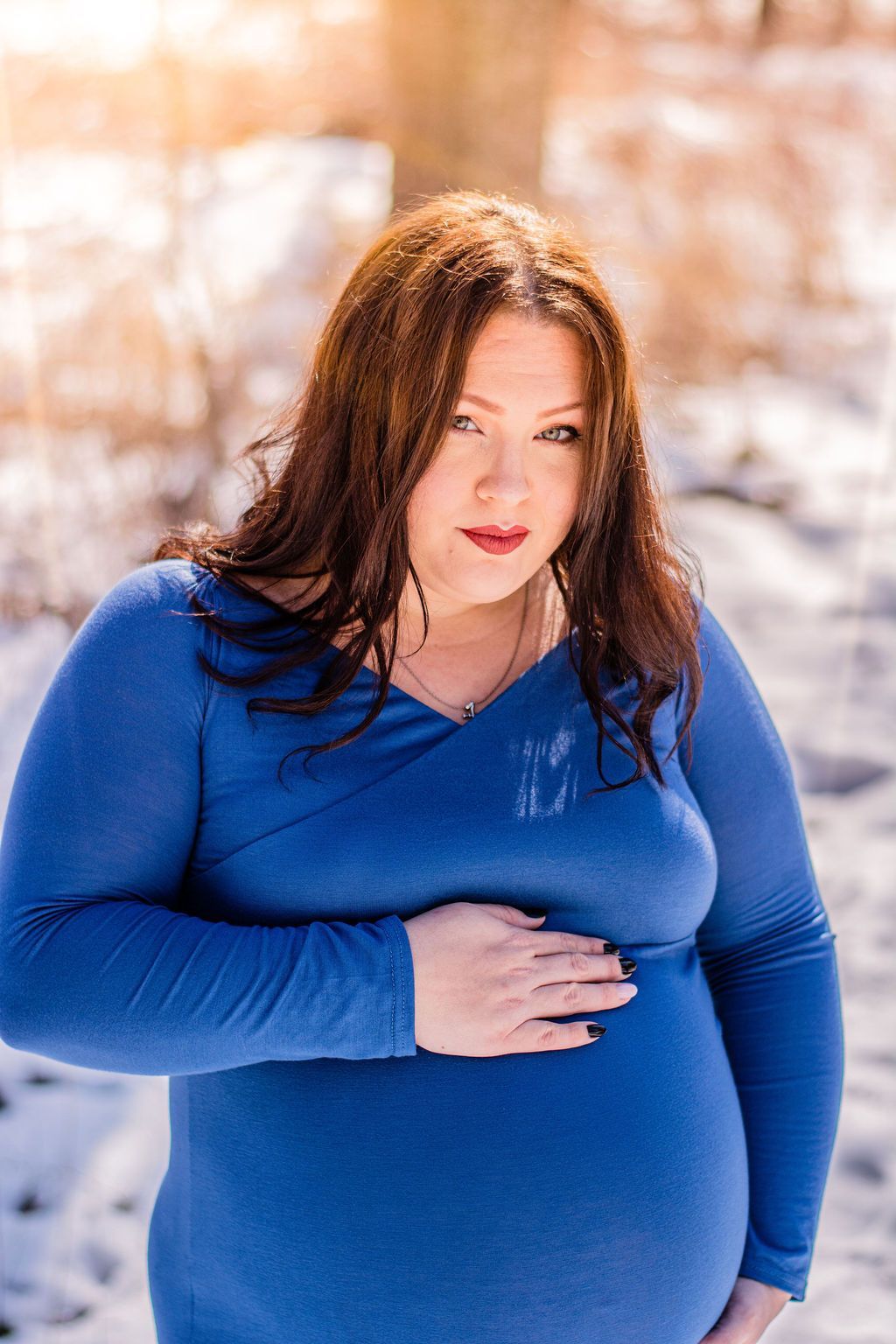 Person in a blue dress standing outdoors in the snow, holding a hand over their belly.